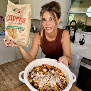 A woman in a kitchen holds a bag of tortilla chips and presents a large bowl of colorful bean and corn salad topped with cheese, smiling at the camera.