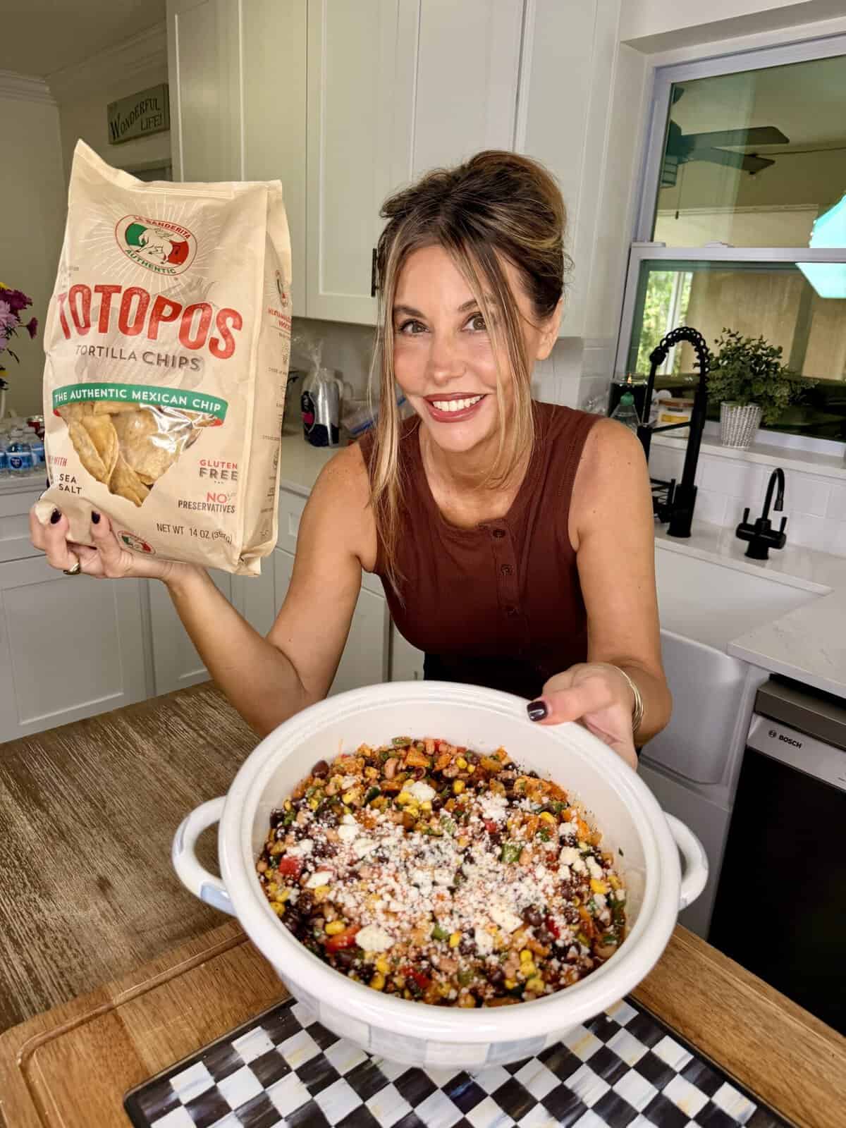 A woman in a kitchen holds a bag of tortilla chips and presents a large bowl of colorful bean and corn salad topped with cheese, smiling at the camera.