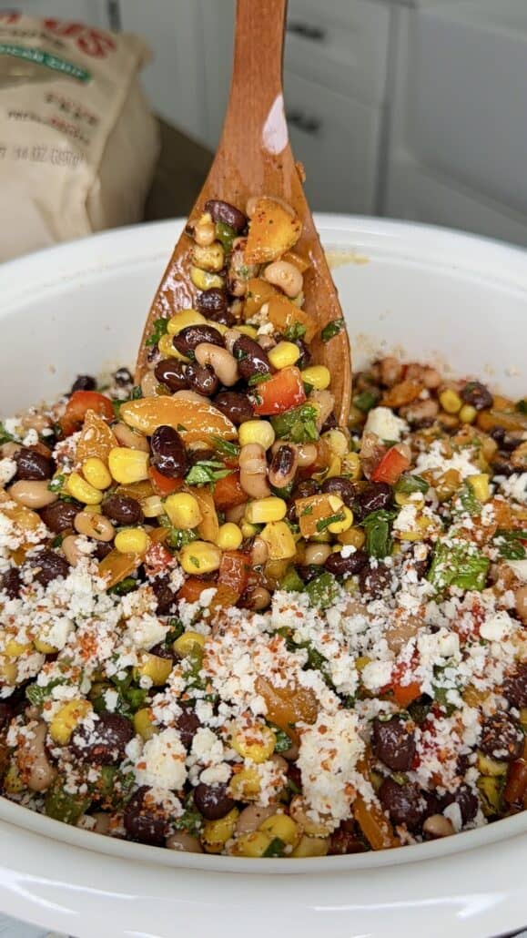 A wooden spoon mixes a colorful salad with corn, black beans, chopped vegetables, herbs, and crumbled cheese in a white bowl. Kitchen counters and a bag are visible in the background.