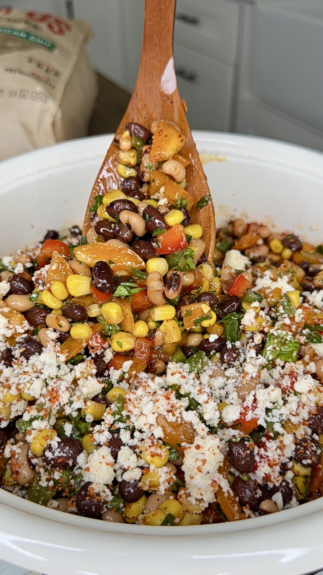 A wooden spoon mixes a colorful salad with corn, black beans, chopped vegetables, herbs, and crumbled cheese in a white bowl. Kitchen counters and a bag are visible in the background.