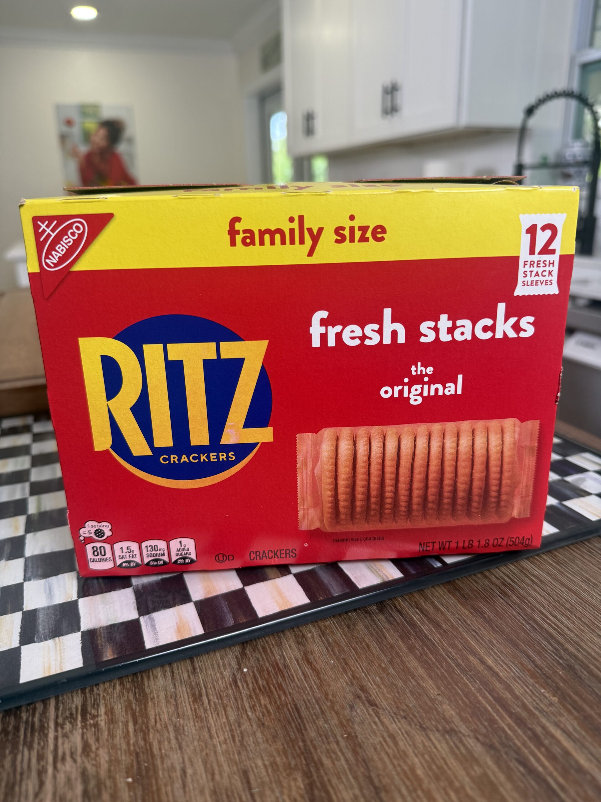 A box of Ritz Crackers family size, labeled fresh stacks the original, containing 12 fresh stack sleeves, sits on a checkered surface in a kitchen setting.