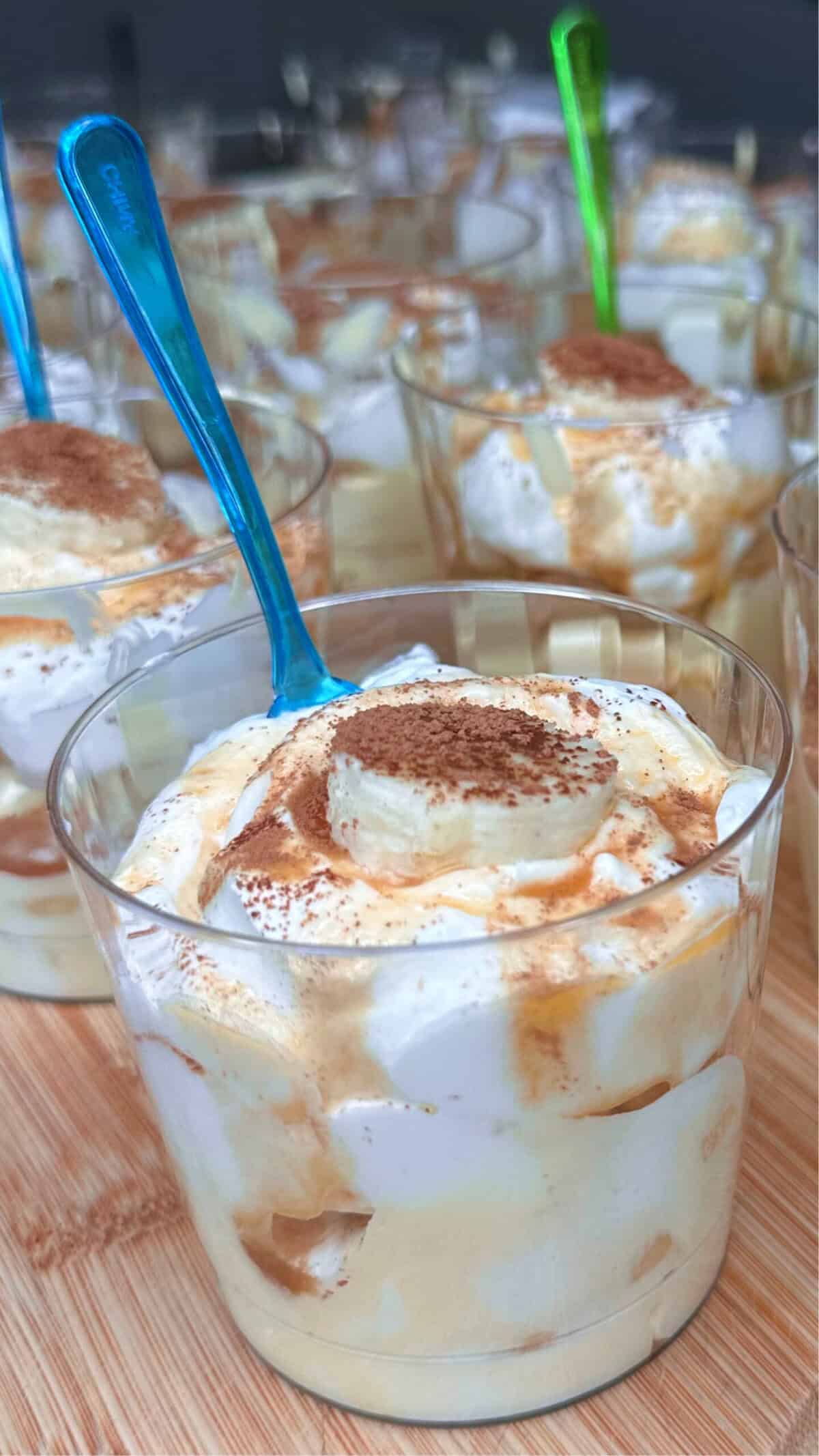 Individual clear cups filled with a creamy dessert, topped with banana slices, whipped cream, and a sprinkle of cocoa powder. Each cup has a colorful plastic spoon and sits on a wooden surface.