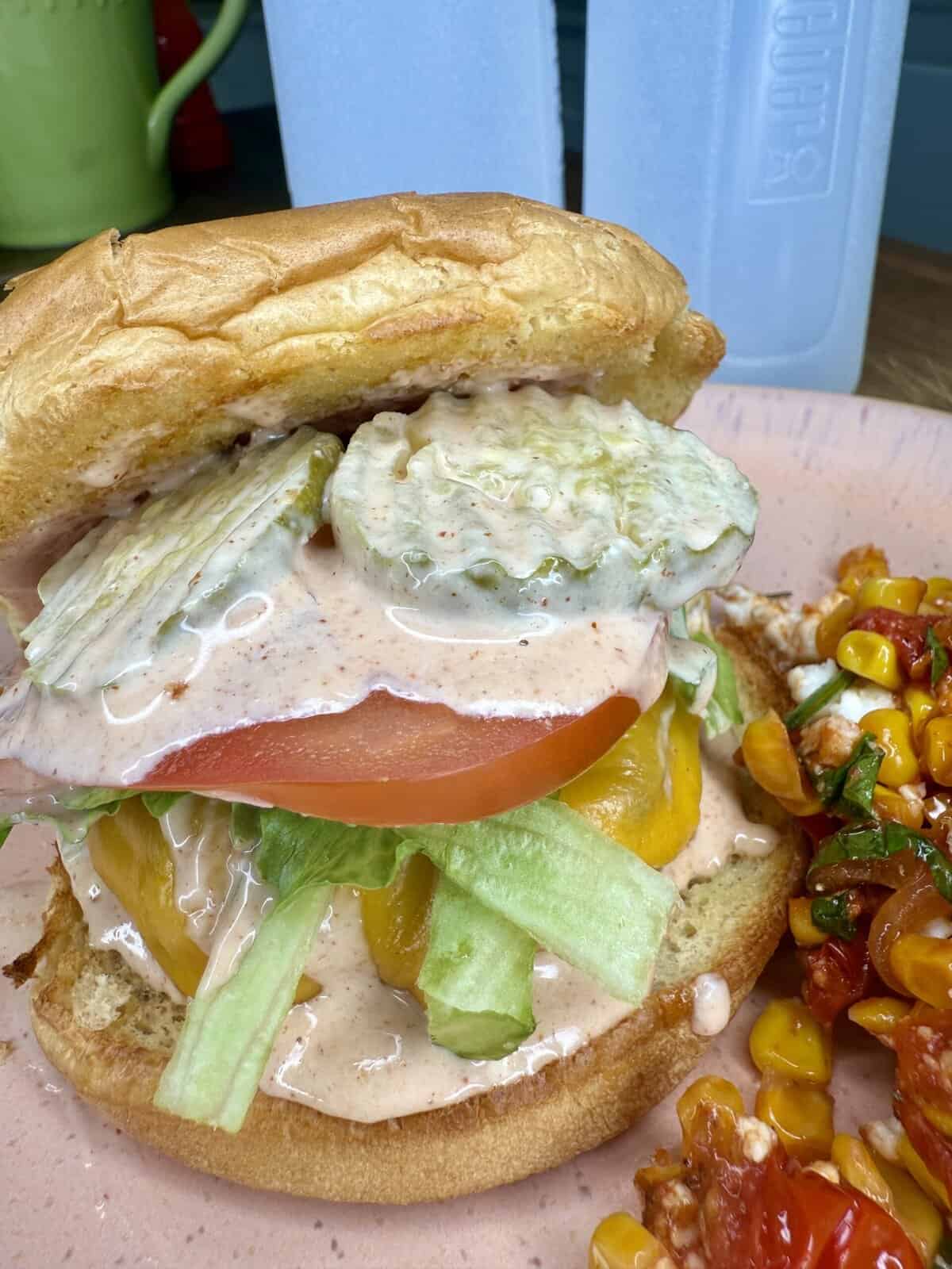 A sandwich with lettuce, tomato, pickles, and creamy sauce in a toasted bun sits on a plate next to a side of corn and tomato salad. Two blue water bottles are in the background.