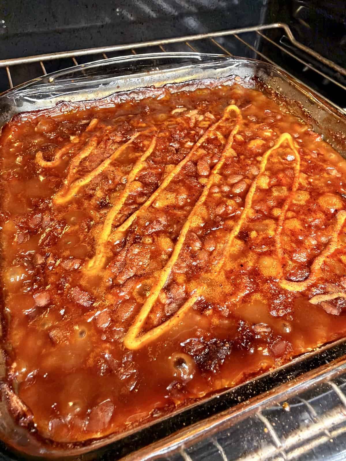A glass baking dish filled with baked beans topped with a drizzle of mustard bakes in an oven, showing a bubbling, golden-brown surface.