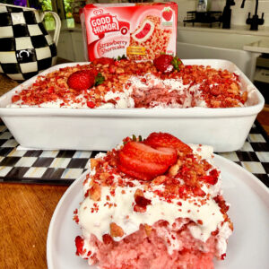A slice of strawberry shortcake with whipped topping and sliced strawberries is on a plate in front of a baking dish of the same dessert. A box of Good Humor Strawberry Shortcake bars is in the background on a checkered tray.