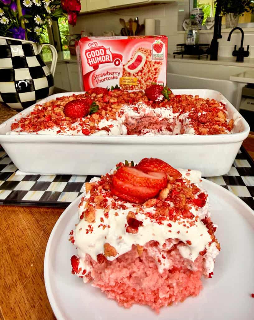 A slice of strawberry shortcake with whipped topping and sliced strawberries is on a plate in front of a baking dish of the same dessert. A box of Good Humor Strawberry Shortcake bars is in the background on a checkered tray.