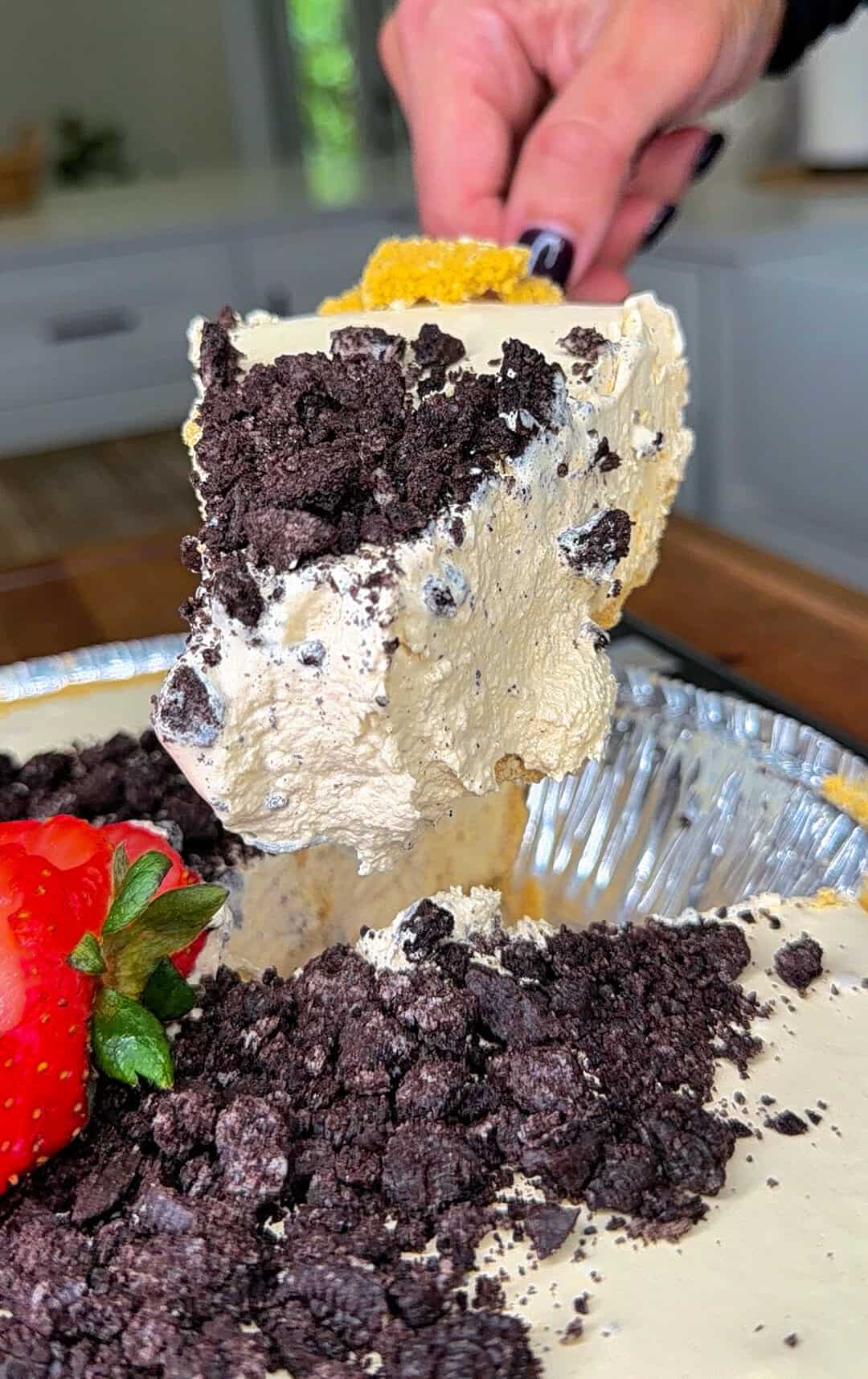 A hand lifts a large, creamy slice of pie topped with crushed chocolate cookies from a pie tin. A fresh strawberry sits beside the pie, and the background shows a kitchen setting.