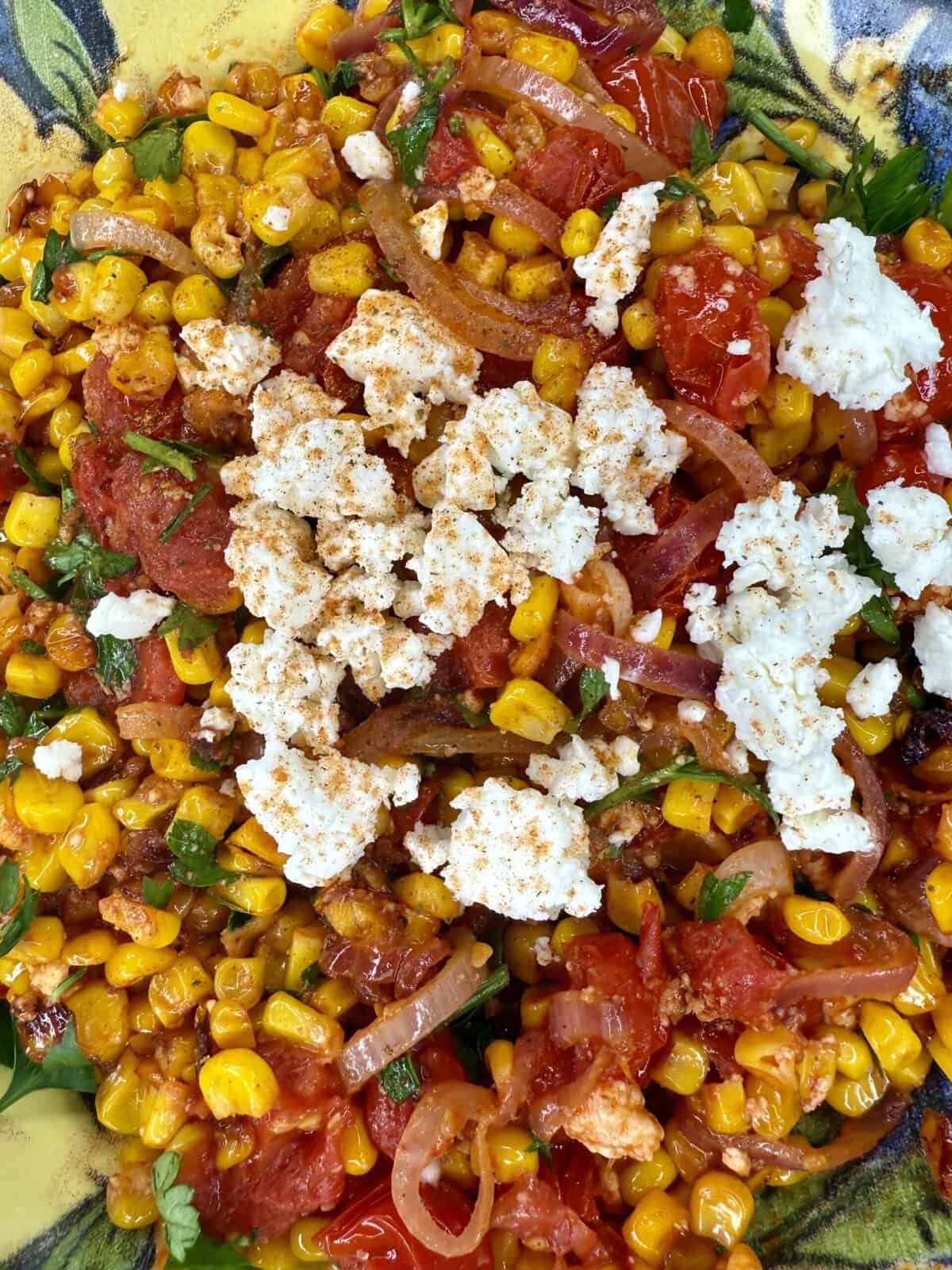 A close-up of a colorful dish featuring roasted corn, sliced red onions, tomato, fresh herbs, and crumbled white cheese, all seasoned with spices and served on a decorative plate.