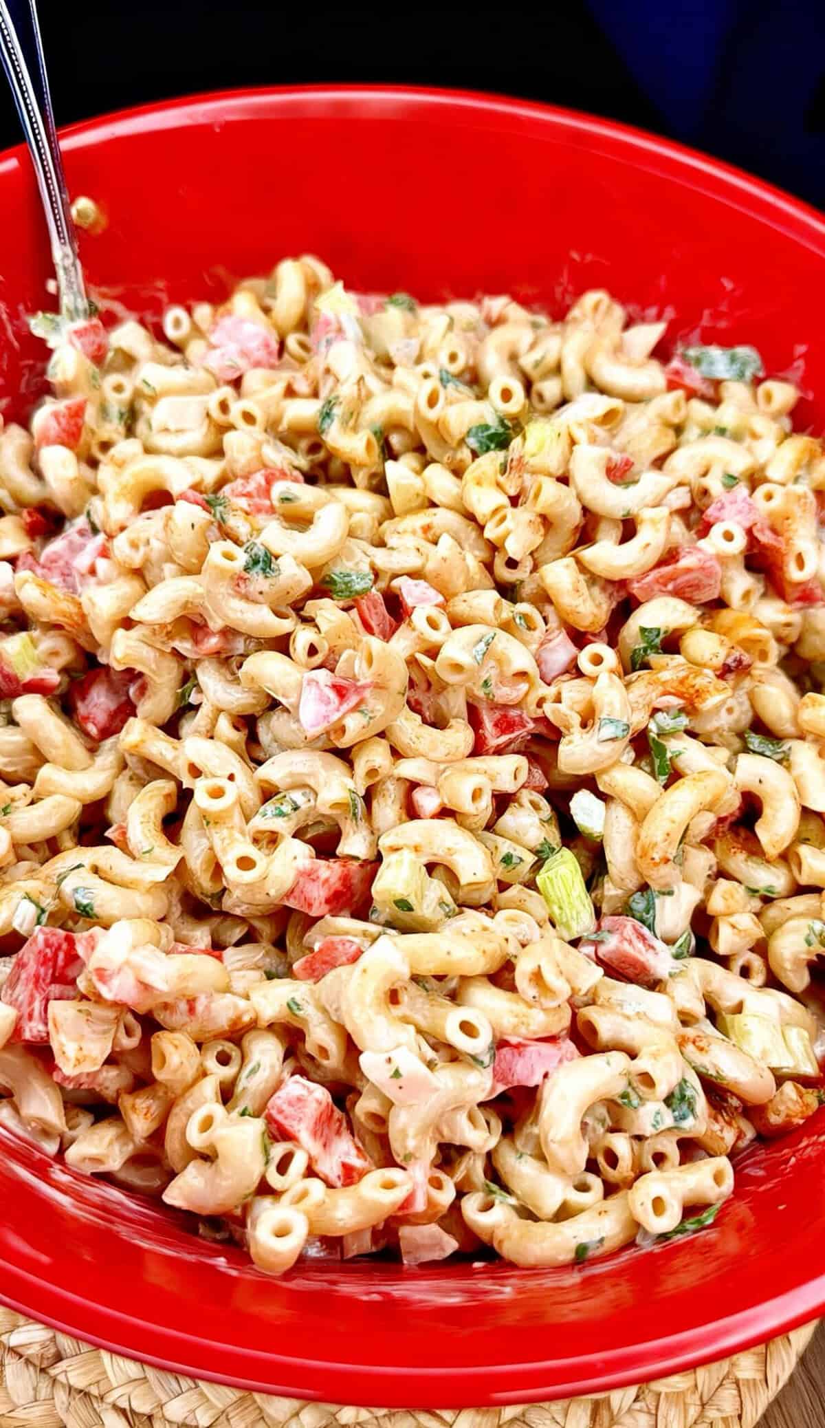 A close-up of creamy macaroni salad with diced red bell peppers, green herbs, and a mayonnaise-based dressing, served in a bright red bowl with a metal spoon.