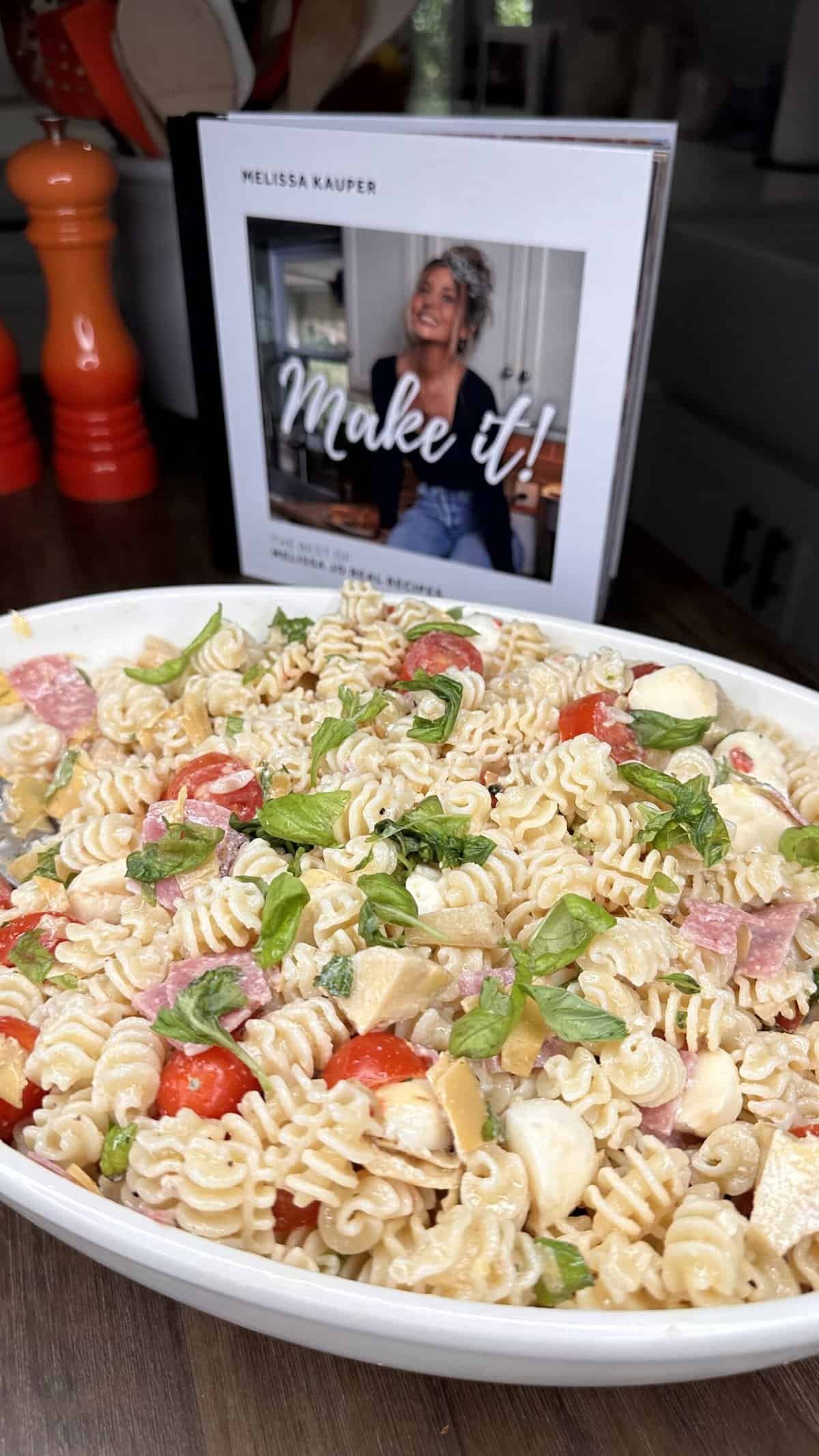A white dish filled with creamy pasta salad, featuring rotini pasta, cherry tomatoes, mozzarella, basil, and salami. In the background is the cookbook Make it! by Melissa Kauper, propped open on a table.