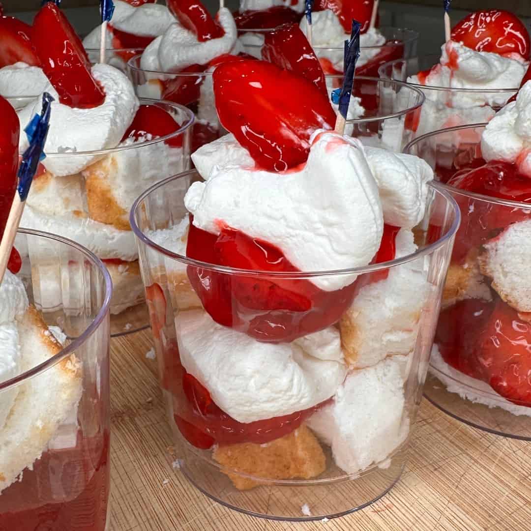 Individual clear cups filled with layers of angel food cake, bright red strawberries in syrup, and whipped cream, all arranged in a dessert parfait style on a wooden surface.