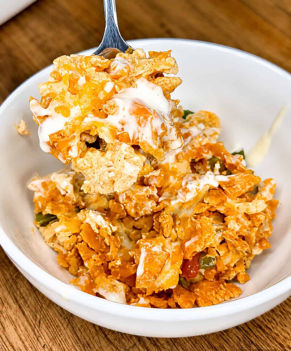 A fork holds a bite of a cheesy, crunchy casserole with crumbled chips and melted cheese above a white bowl filled with the dish.