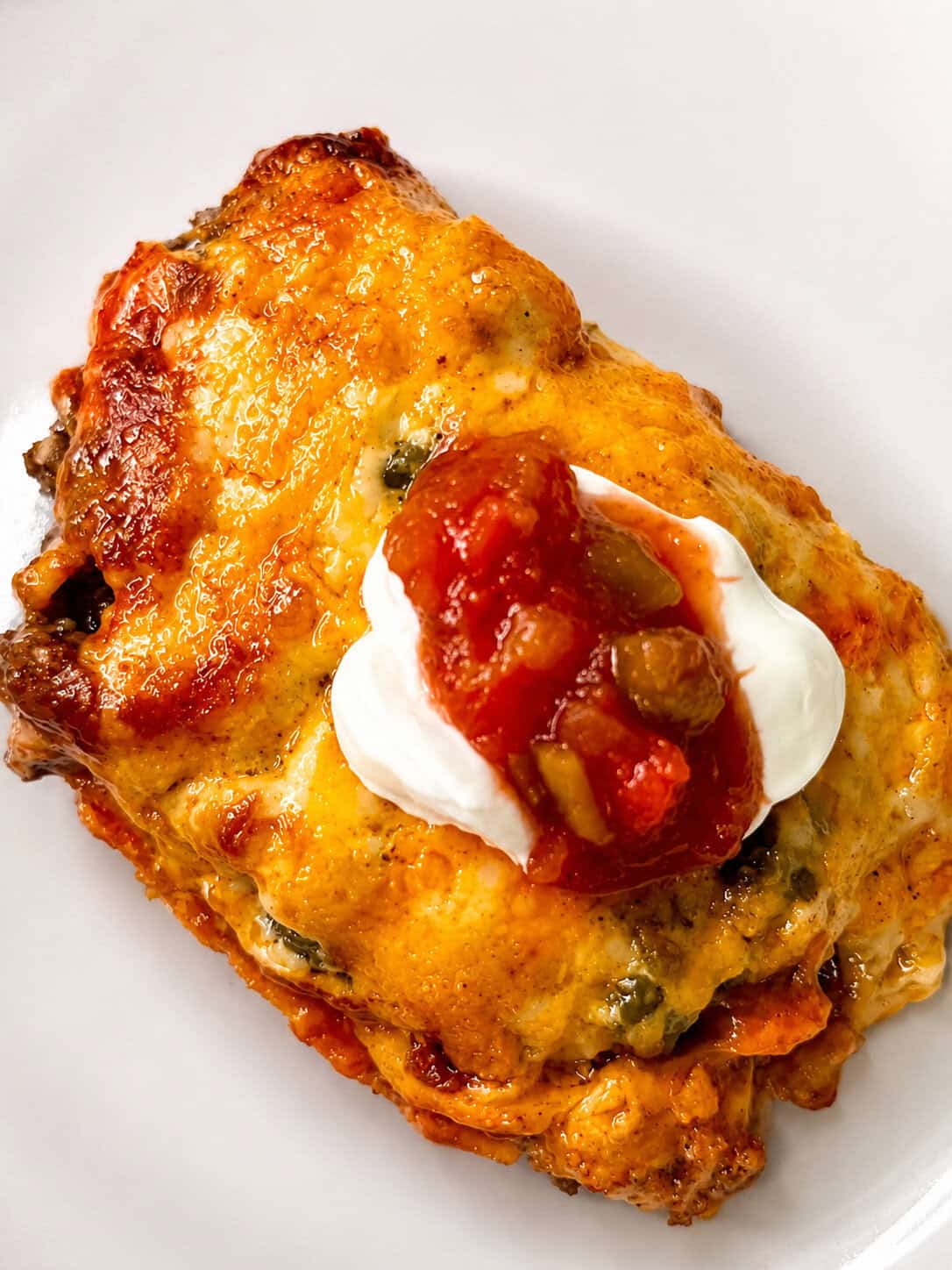 A cheesy baked casserole topped with a dollop of sour cream and chunky salsa sits on a white plate.