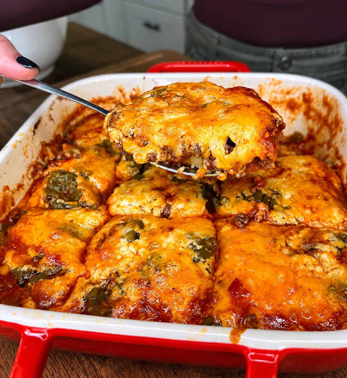A person serves a cheesy baked casserole from a red baking dish, revealing gooey melted cheese and a tomato-based sauce with green peppers.