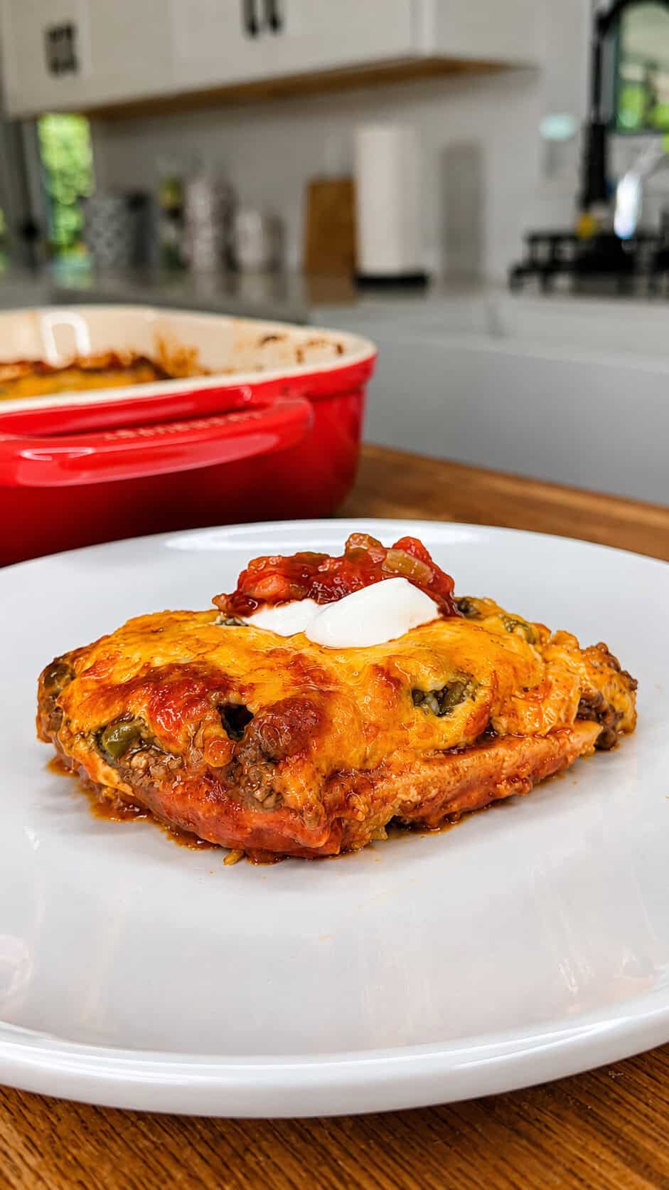 A slice of cheesy casserole topped with sour cream and salsa on a white plate, with a red baking dish and modern kitchen in the background.
