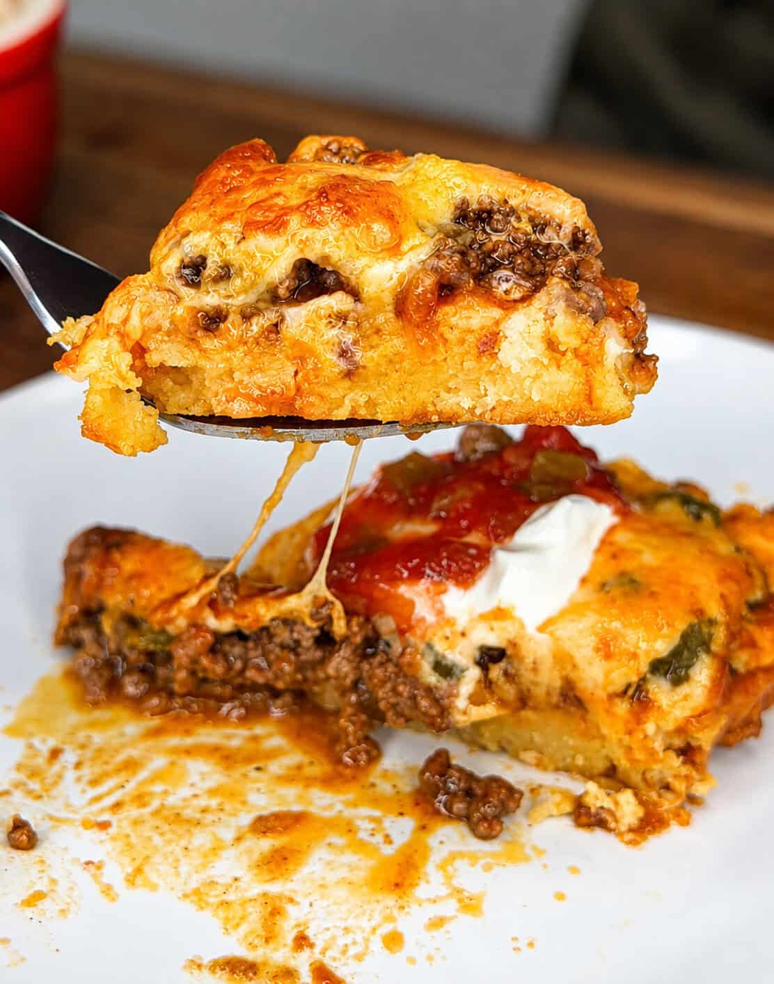 A close-up of a cheesy, baked casserole with ground beef, topped with salsa and sour cream, lifted by a fork over a white plate with more casserole and sauce.
