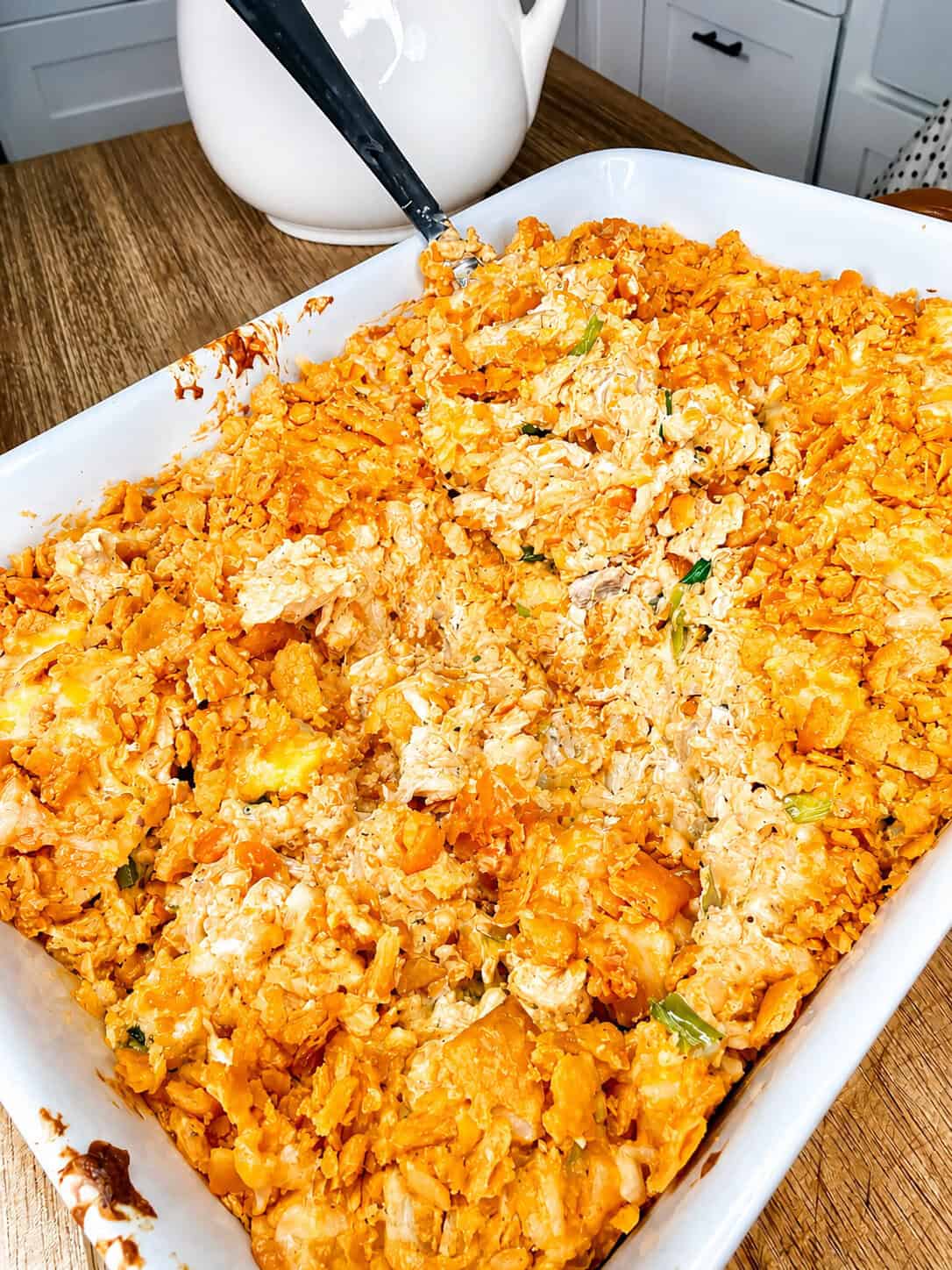A casserole dish filled with a cheesy, crunchy-topped baked casserole, possibly containing chicken and vegetables, sits on a wooden table with a serving spoon inside.