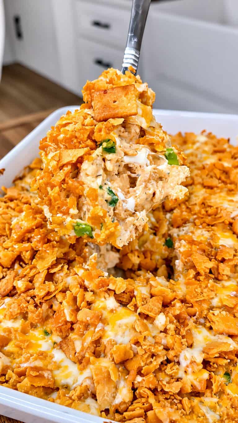 A spoon lifts a cheesy, golden-brown casserole topped with crushed crackers and green onions from a white baking dish.