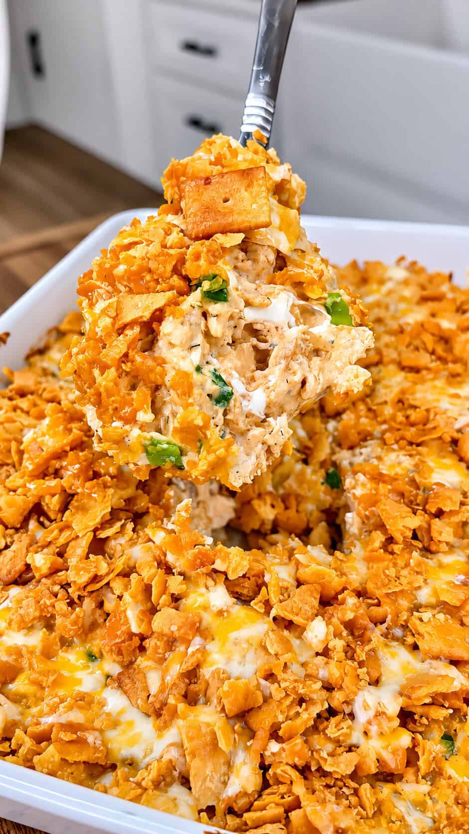 A spoon lifts a cheesy, golden-brown casserole topped with crushed crackers and green onions from a white baking dish.