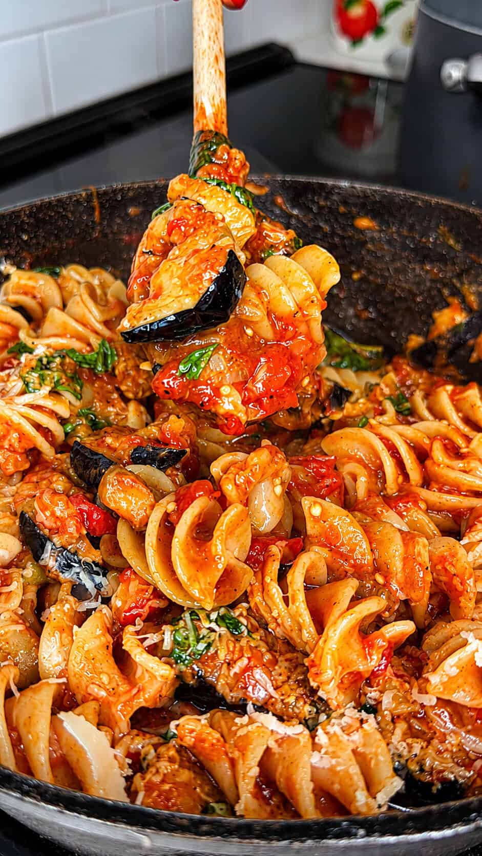 A close-up of a skillet filled with rotini pasta, tomato sauce, eggplant, and fresh basil, with a wooden spoon lifting a serving.
