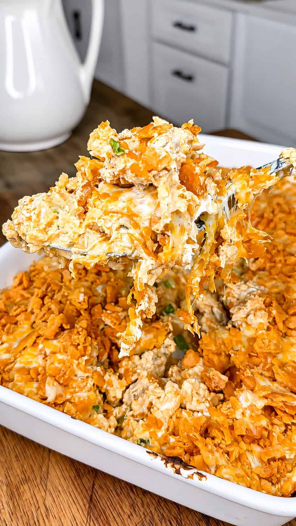 A serving spoon lifts a cheesy, creamy casserole topped with crushed, orange chips from a white baking dish in a kitchen setting.