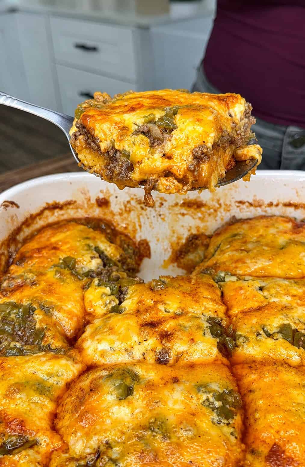 A metal spatula lifts a cheesy, beef-filled casserole piece from a baking dish, showing melted cheese, ground beef, and green peppers. The casserole is cut into squares.