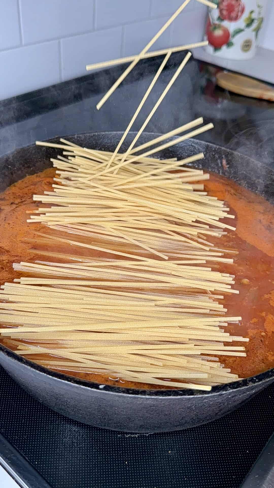 Uncooked spaghetti being added to a black skillet filled with red tomato sauce on a stove, with some pasta pieces suspended in midair above the pan.