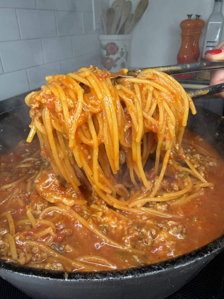 Tongs lifting One Pan Viral Spaghetti with meat sauce from a steaming pot. The sauce is thick and red with visible ground meat and tomatoes. White tiles, utensils, and orange pepper grinders are seen in the kitchen background.