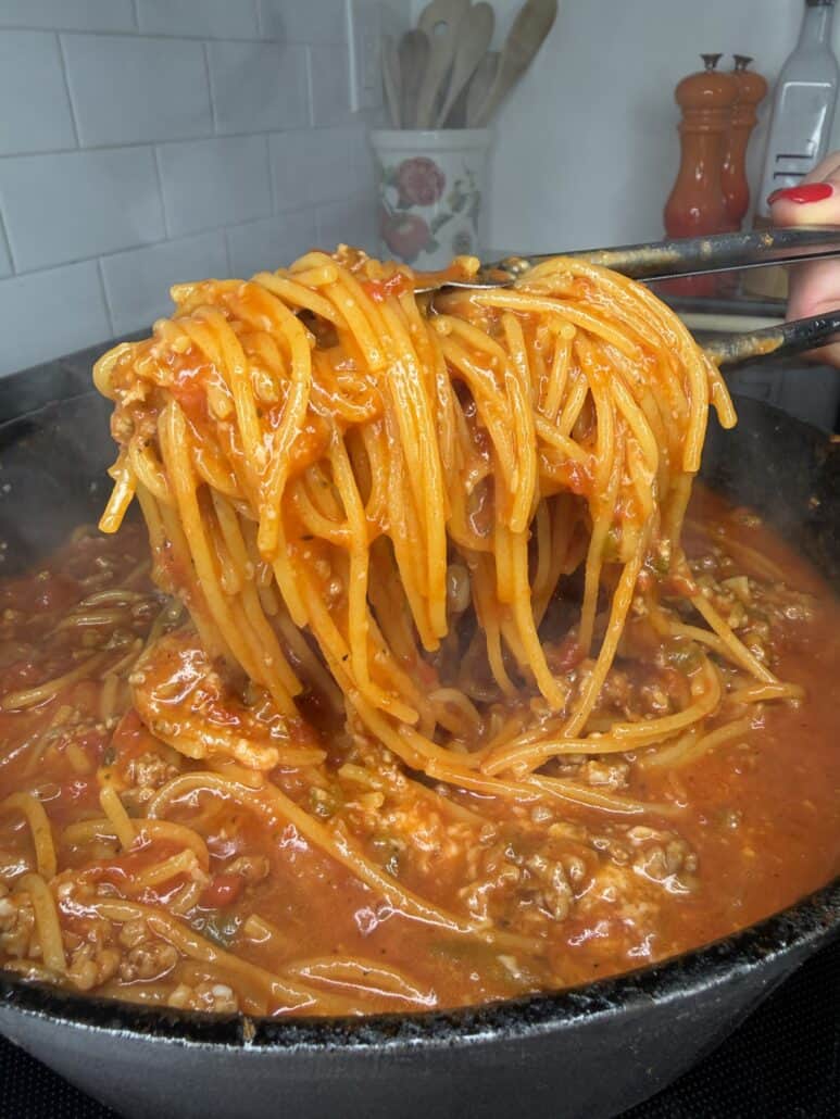 Tongs lifting One Pan Viral Spaghetti with meat sauce from a steaming pot. The sauce is thick and red with visible ground meat and tomatoes. White tiles, utensils, and orange pepper grinders are seen in the kitchen background.