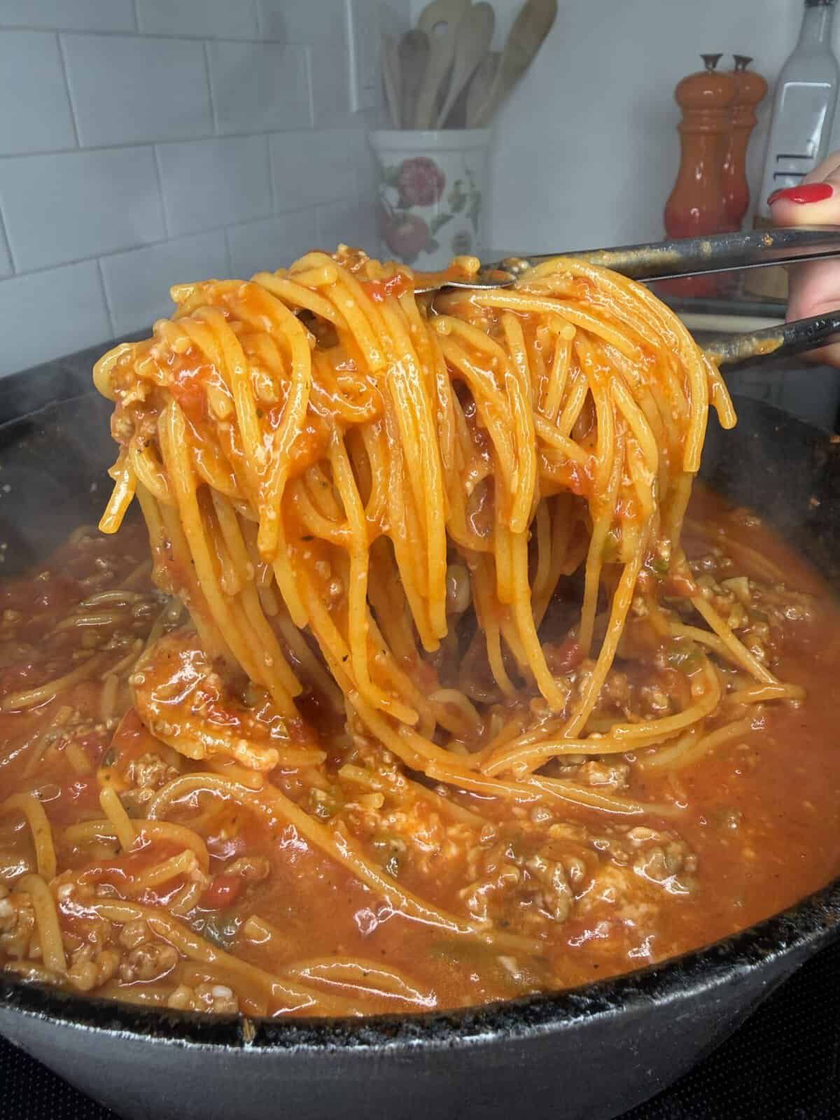 A close-up of spaghetti in tomato meat sauce being lifted from a pot with tongs. The dish is steaming and appears freshly made. Kitchen utensils and spice containers are visible in the background.