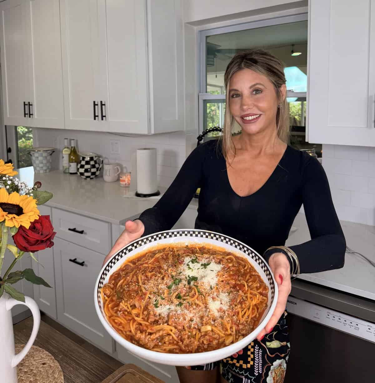 A woman in a kitchen smiles while holding a large plate of spaghetti topped with sauce, cheese, and herbs. A vase with sunflowers sits on the counter beside her.