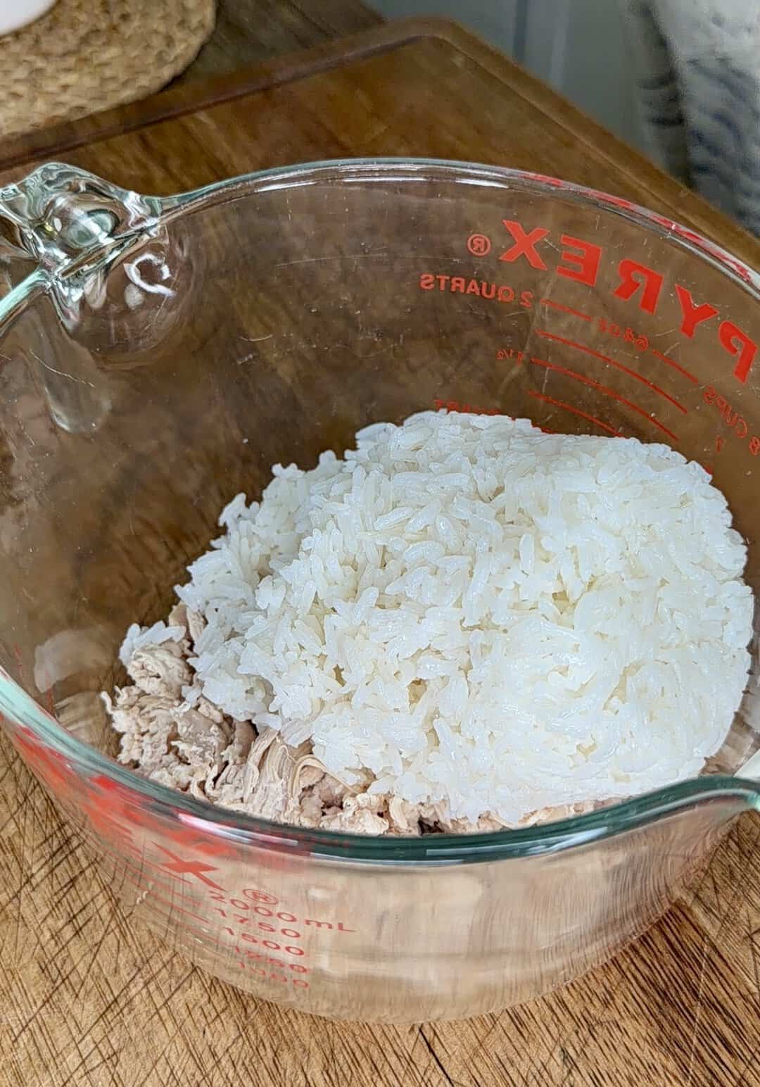 A glass Pyrex measuring cup on a wooden surface contains cooked white rice and shredded meat, ready to be mixed.