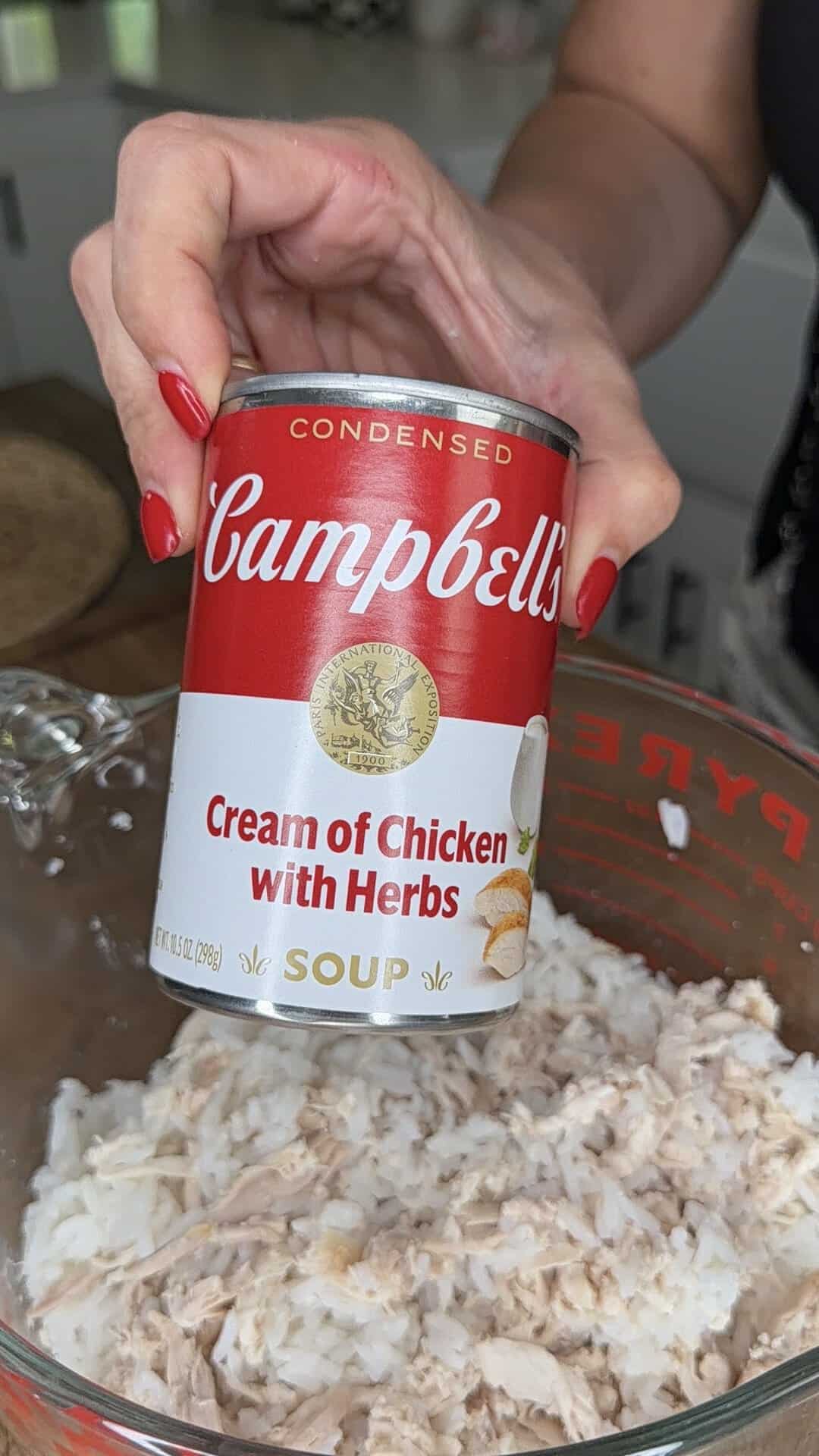A hand with red nails holds a can of Campbell’s Cream of Chicken with Herbs soup over a glass bowl filled with shredded chicken.