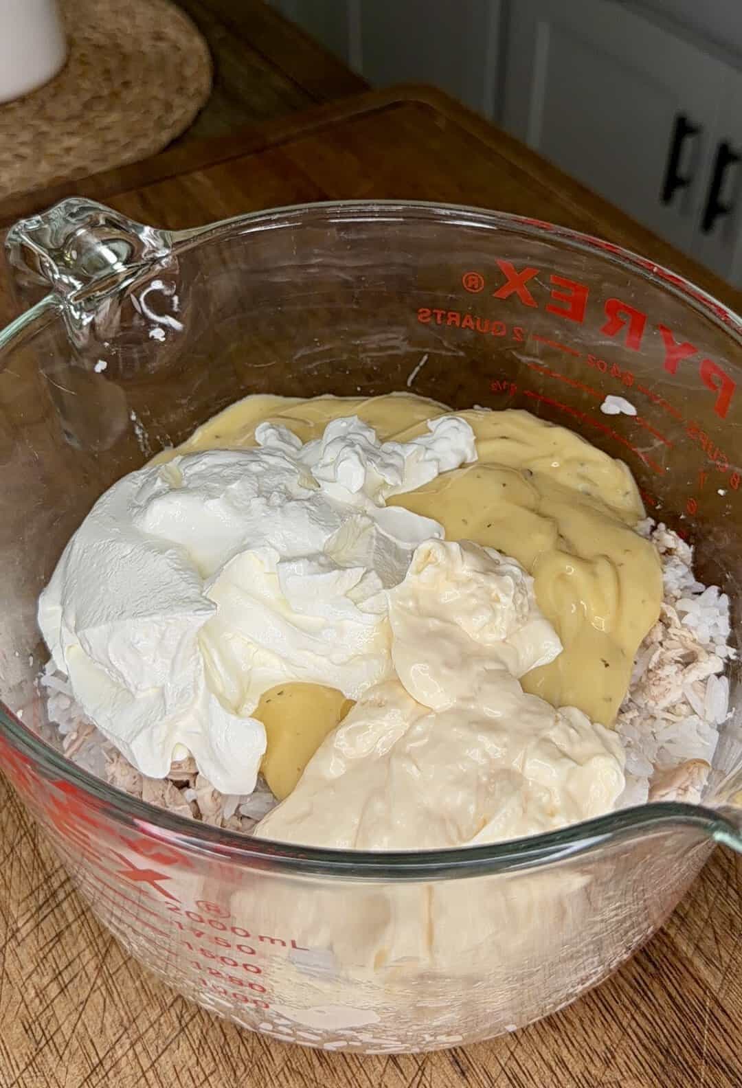 A glass measuring cup filled with shredded chicken, sour cream, cream of chicken soup, and mayonnaise on a wooden countertop.