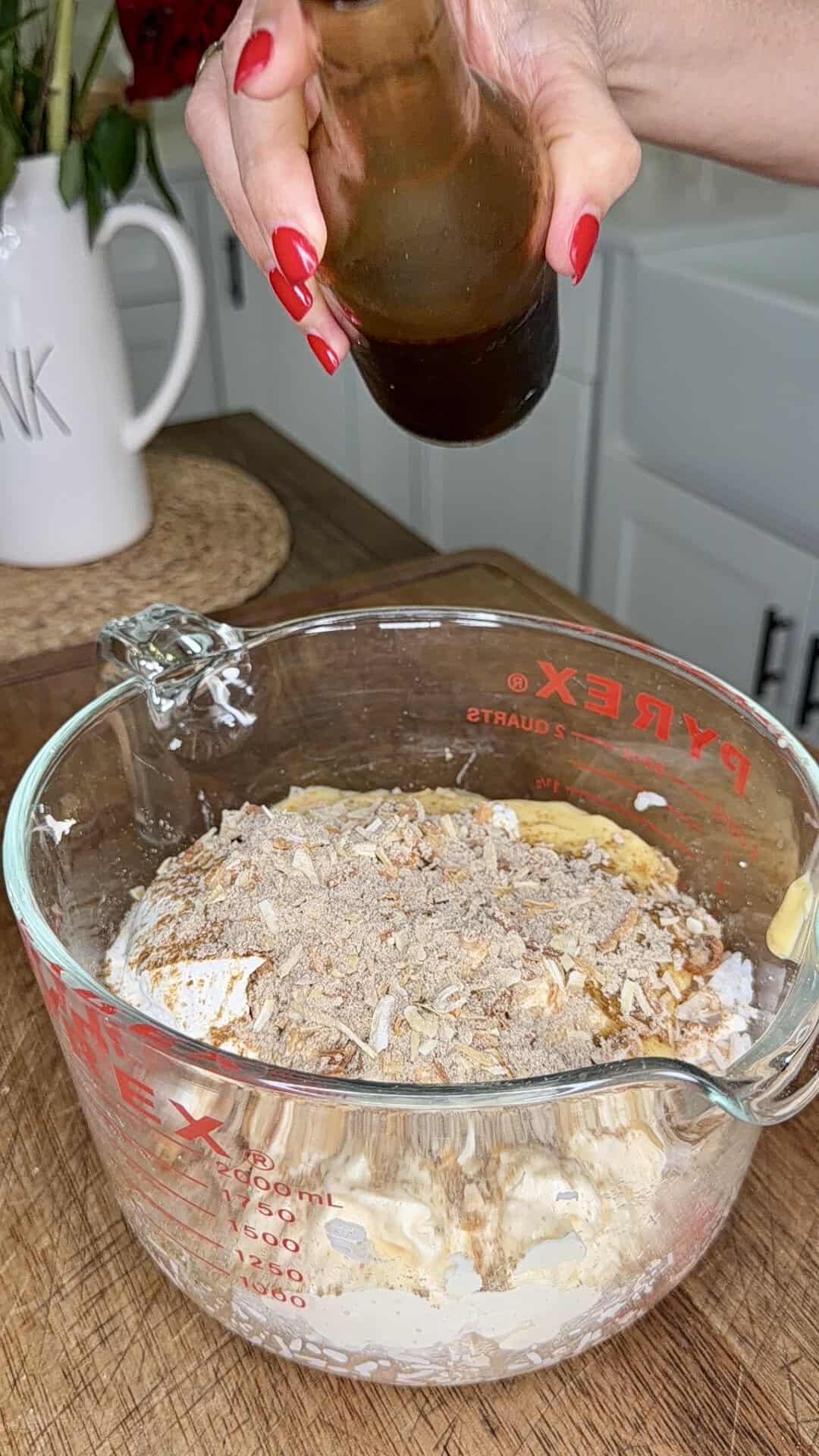 A hand with red nails holds a bottle above a glass mixing bowl filled with ingredients, including crushed crackers, on a wooden surface in a kitchen.