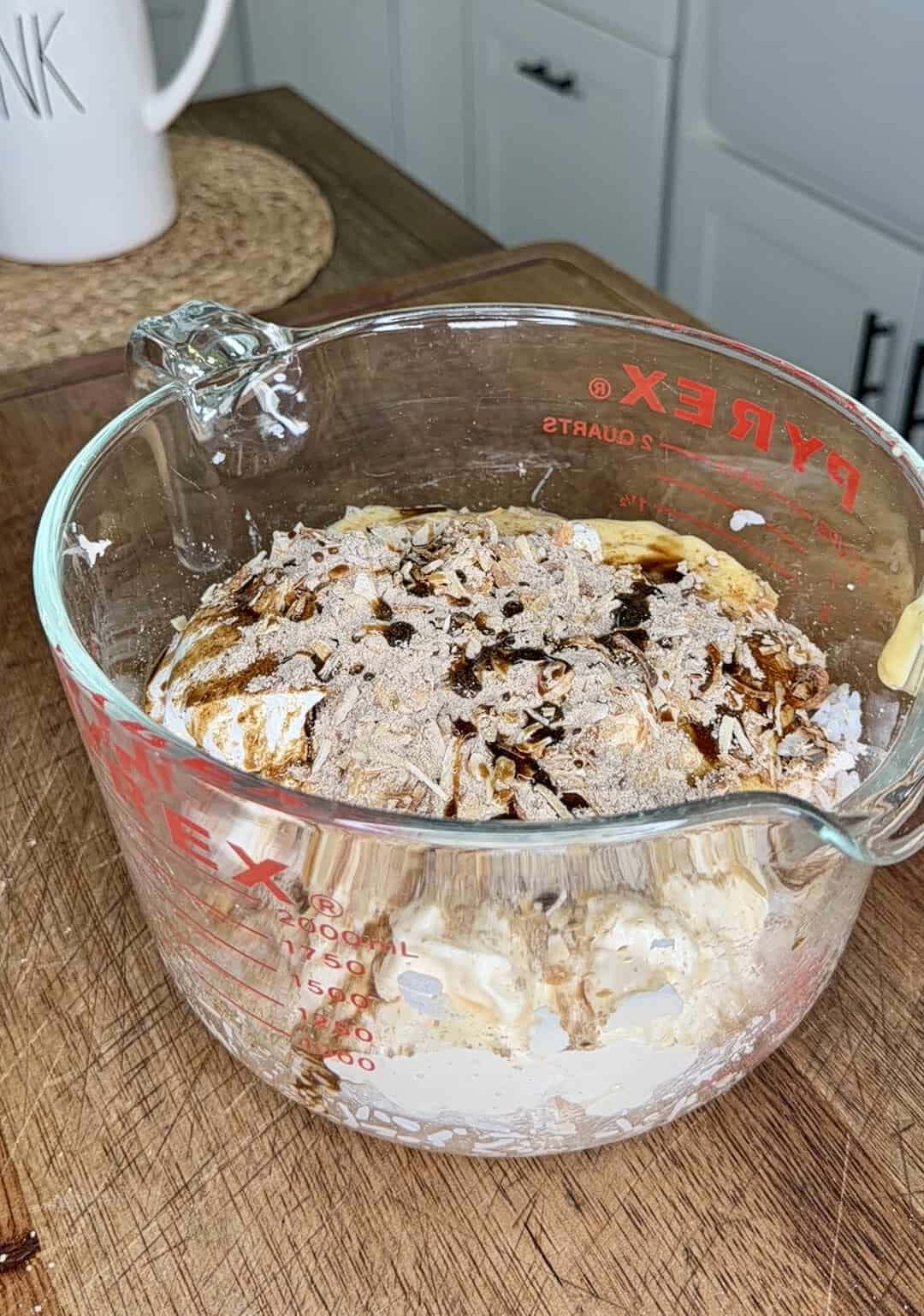A glass measuring cup on a wooden surface contains whipped cream, brown spices, and vanilla, ready to be mixed in a kitchen setting.