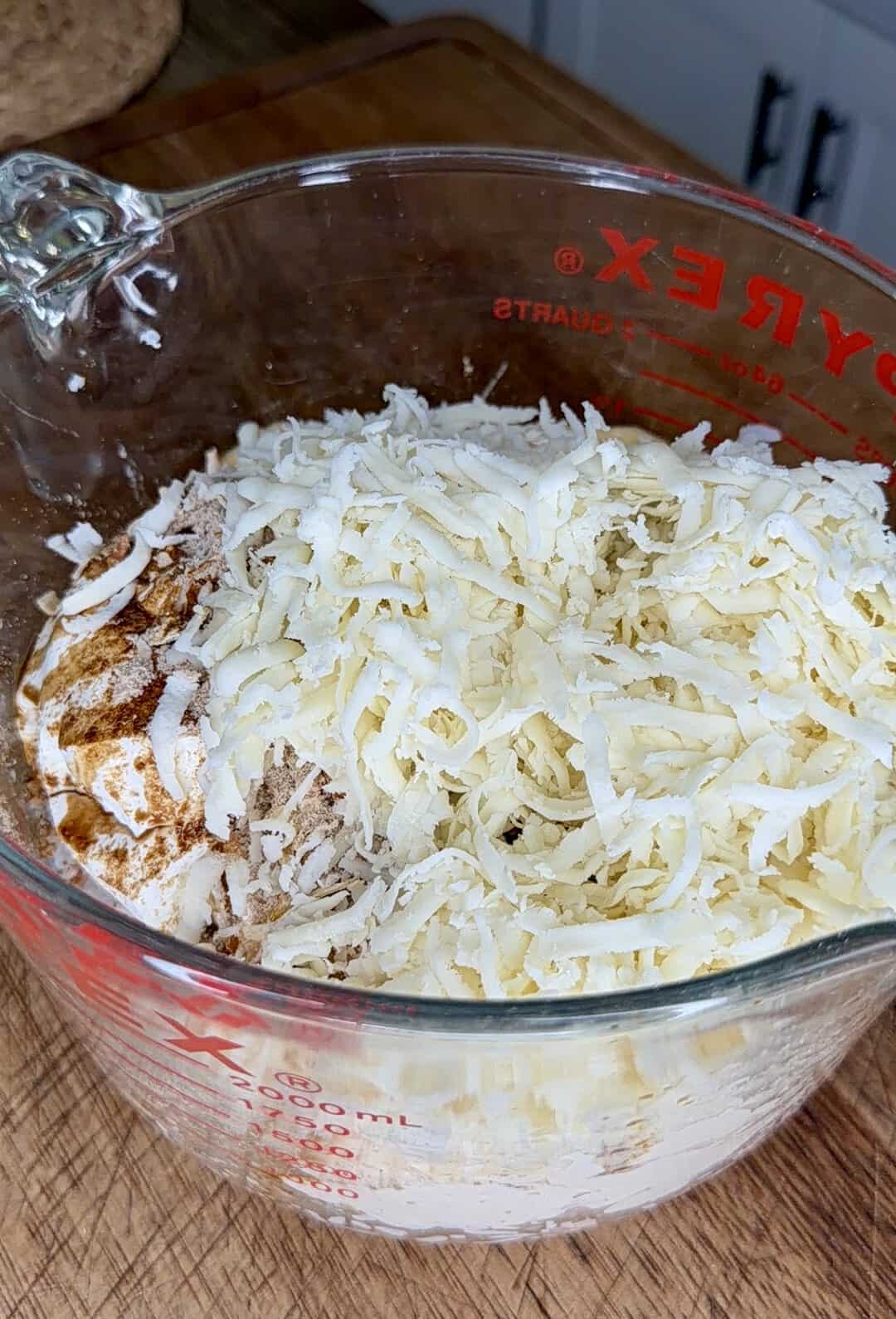 A glass measuring cup filled with shredded cheese, flour, and seasonings sits on a wooden countertop in a kitchen.