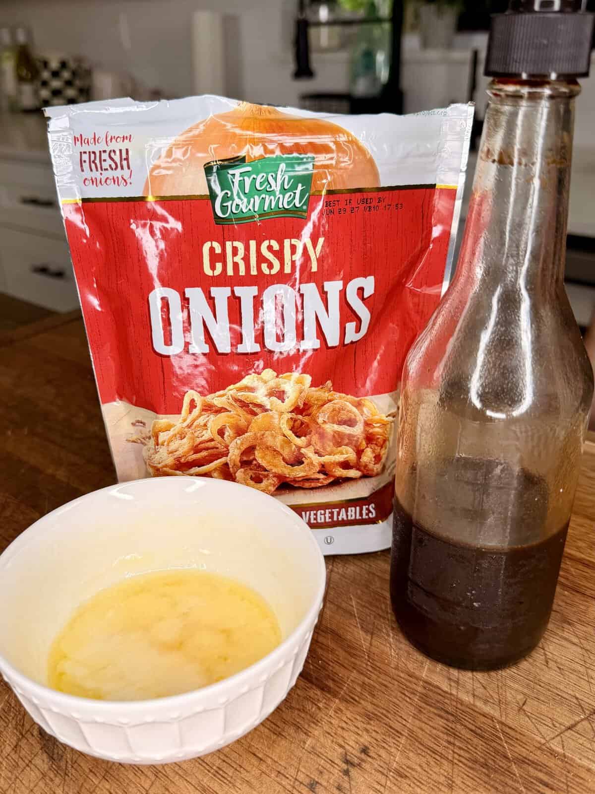 A bag of Fresh Gourmet crispy onions, a brown bottle, and a white bowl with a yellow liquid sit on a wooden countertop in a kitchen.