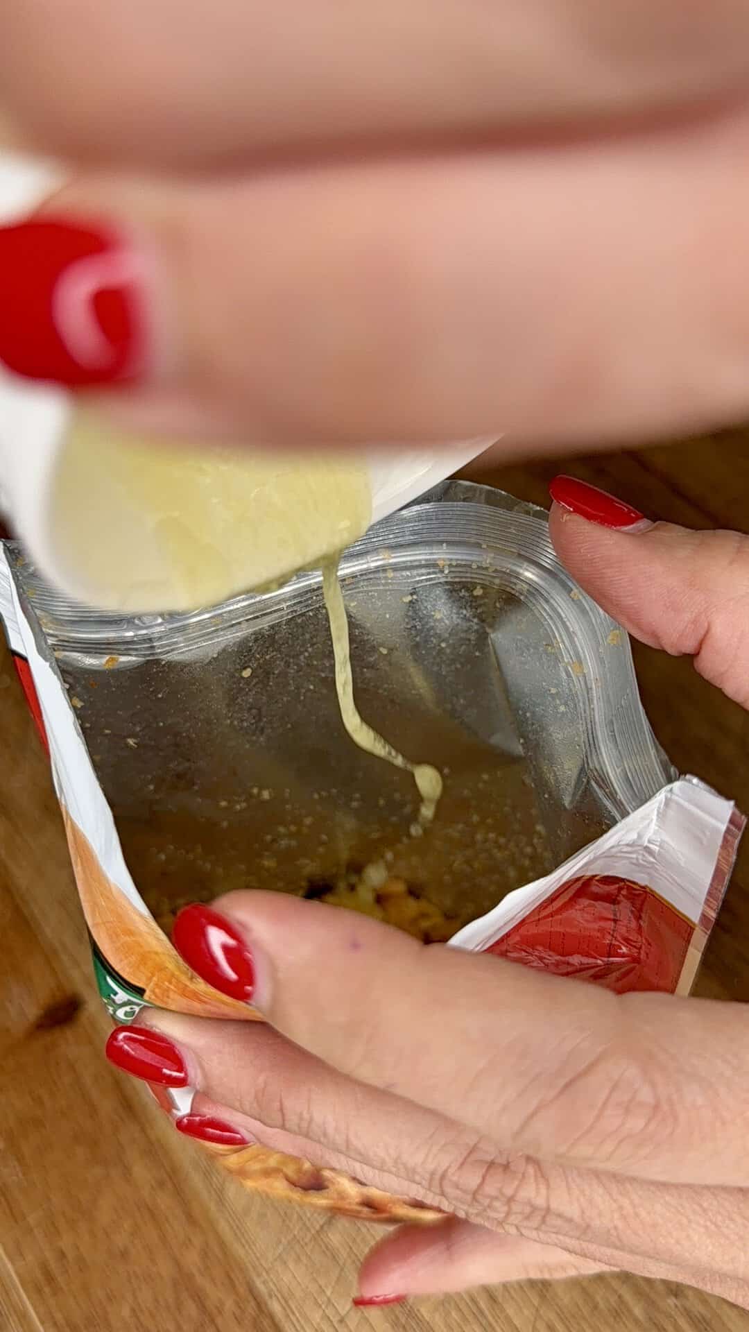 A person with red nail polish pours sauce from a small packet into an opened bag of snacks on a wooden surface.
