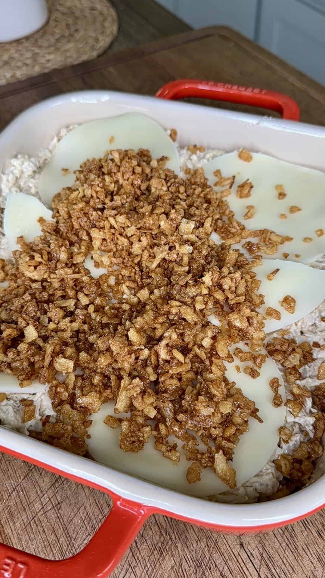 A red baking dish filled with oats, topped with slices of cheese and a layer of crispy fried onions, sits on a wooden countertop.