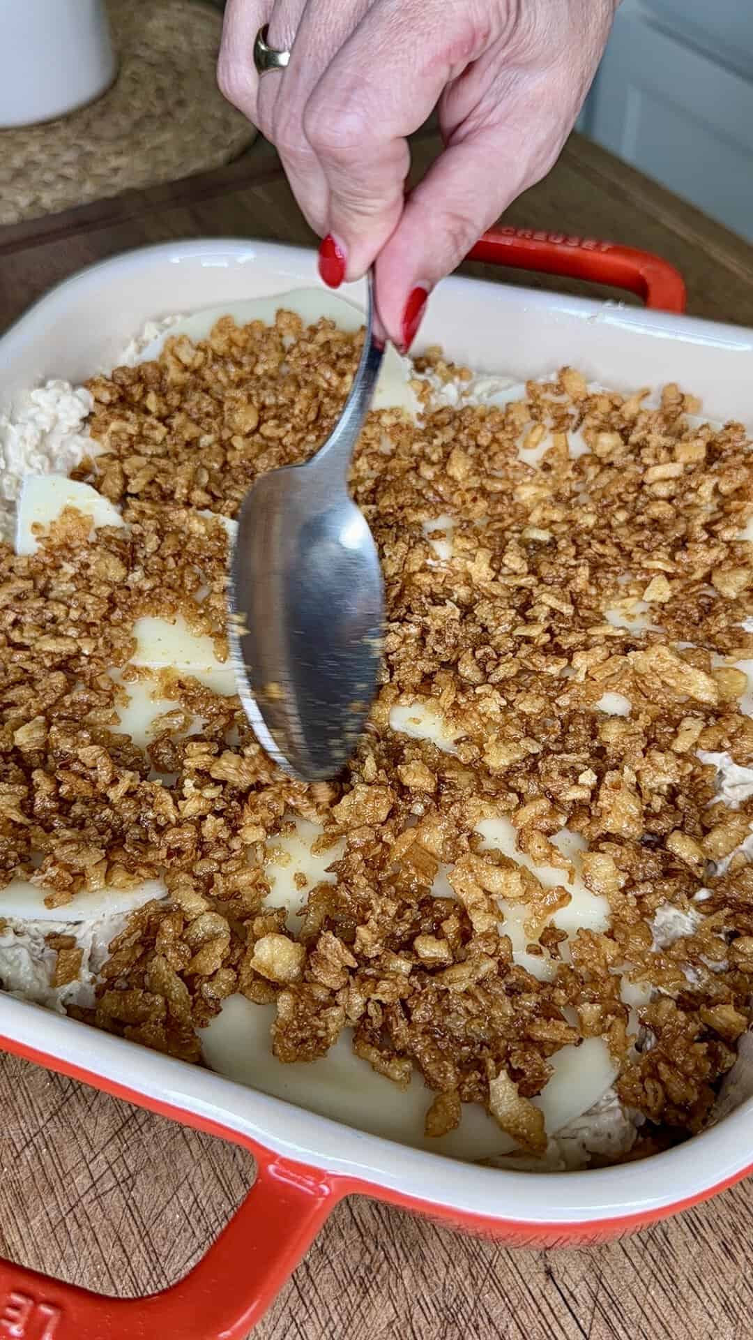 A hand holds a spoon, spreading crispy brown topping over a casserole in a red baking dish, with visible layers of sliced cheese and rice underneath.