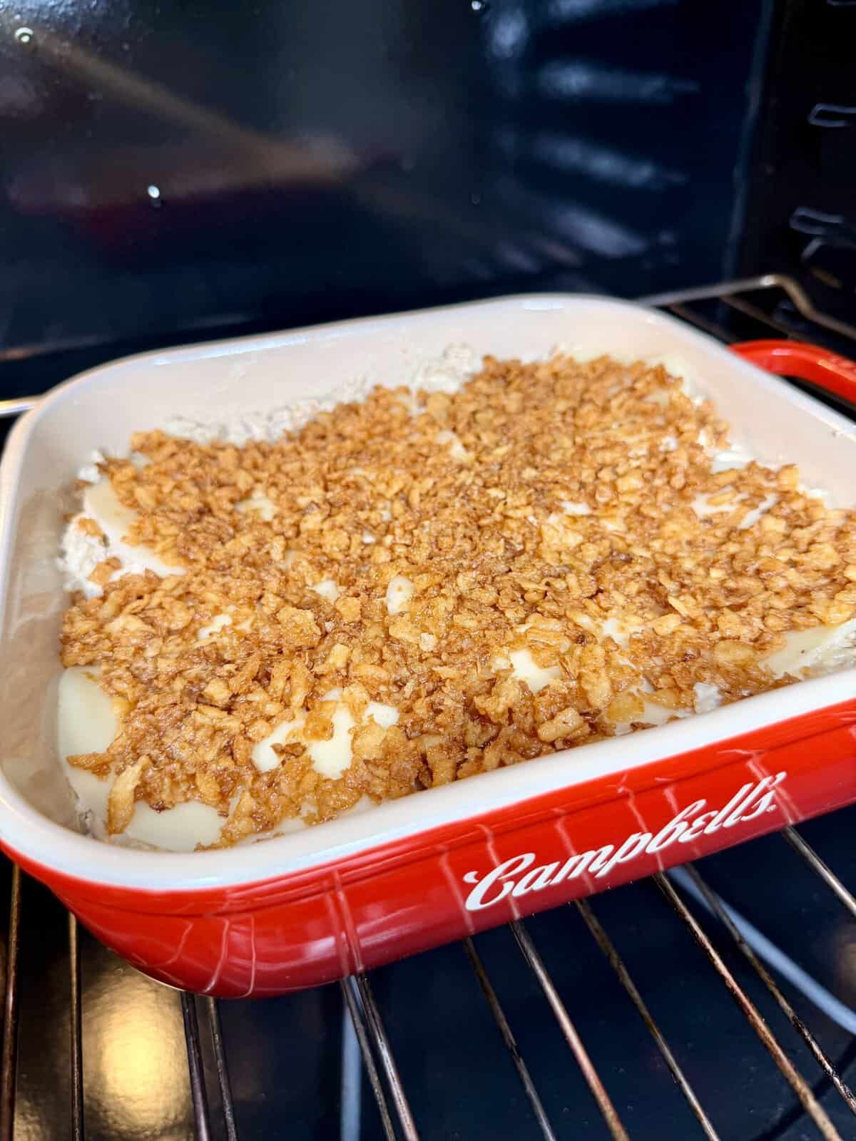 A red casserole dish labeled Campbells sits in an oven, filled with a creamy mixture topped with a layer of crispy, golden brown fried onions.