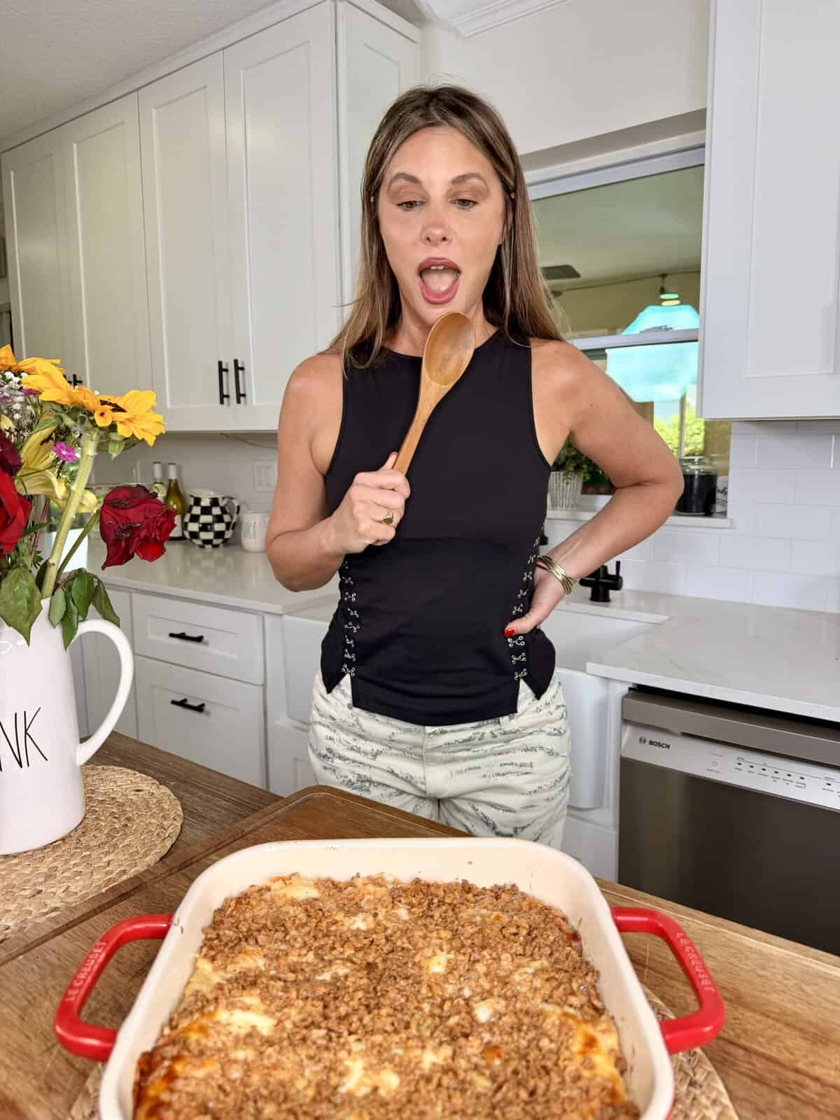 A woman in a black top holds a wooden spoon and looks excitedly at a casserole dish with a crumb topping on a kitchen counter.