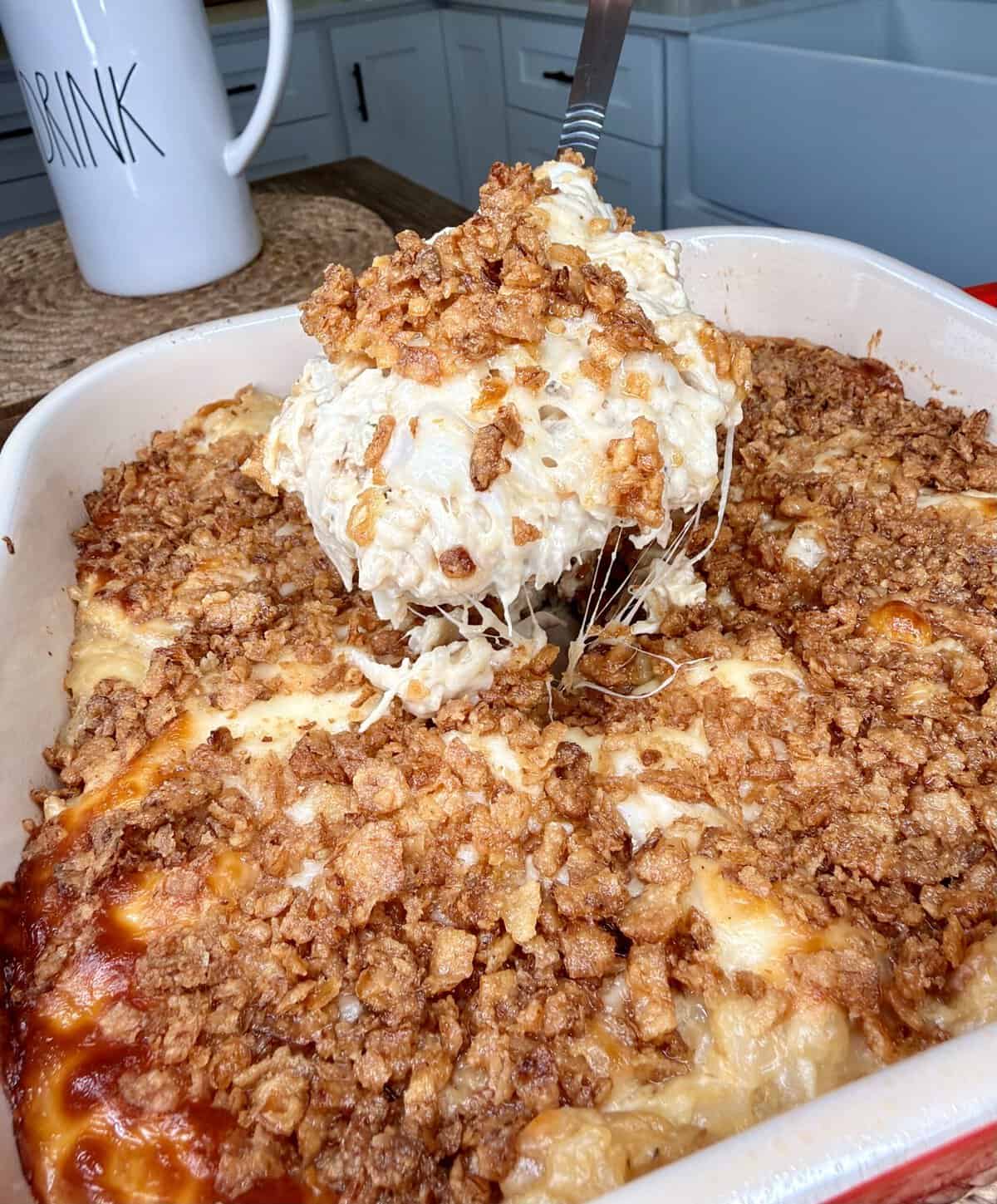 A cheesy casserole topped with a golden, crunchy layer is being served from a baking dish in a kitchen. A mug labeled “DRINK” is in the background on a woven mat.