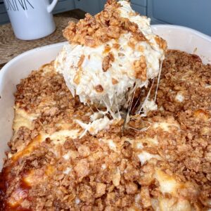 A cheesy casserole with a crunchy topping is being scooped from a baking dish with a spatula. The dish sits on a kitchen counter next to a large white mug.