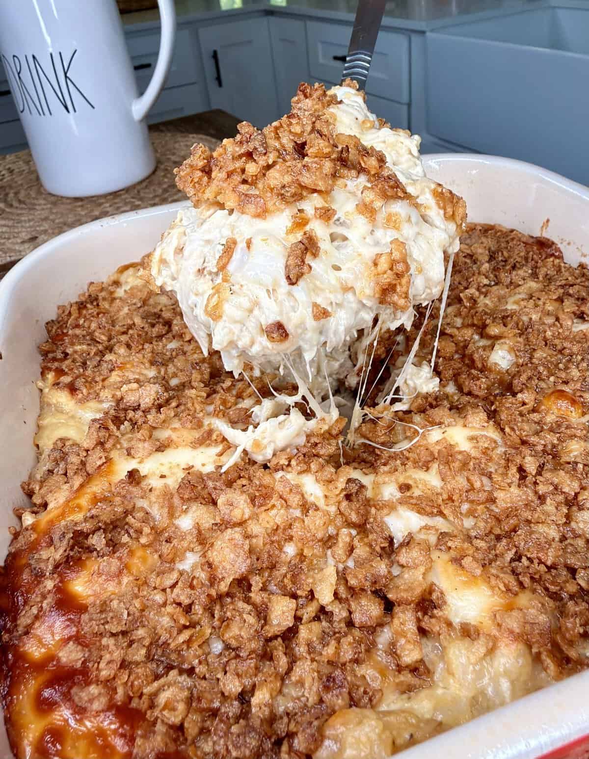 A cheesy casserole with a crunchy topping is being scooped from a baking dish with a spatula. The dish sits on a kitchen counter next to a large white mug.