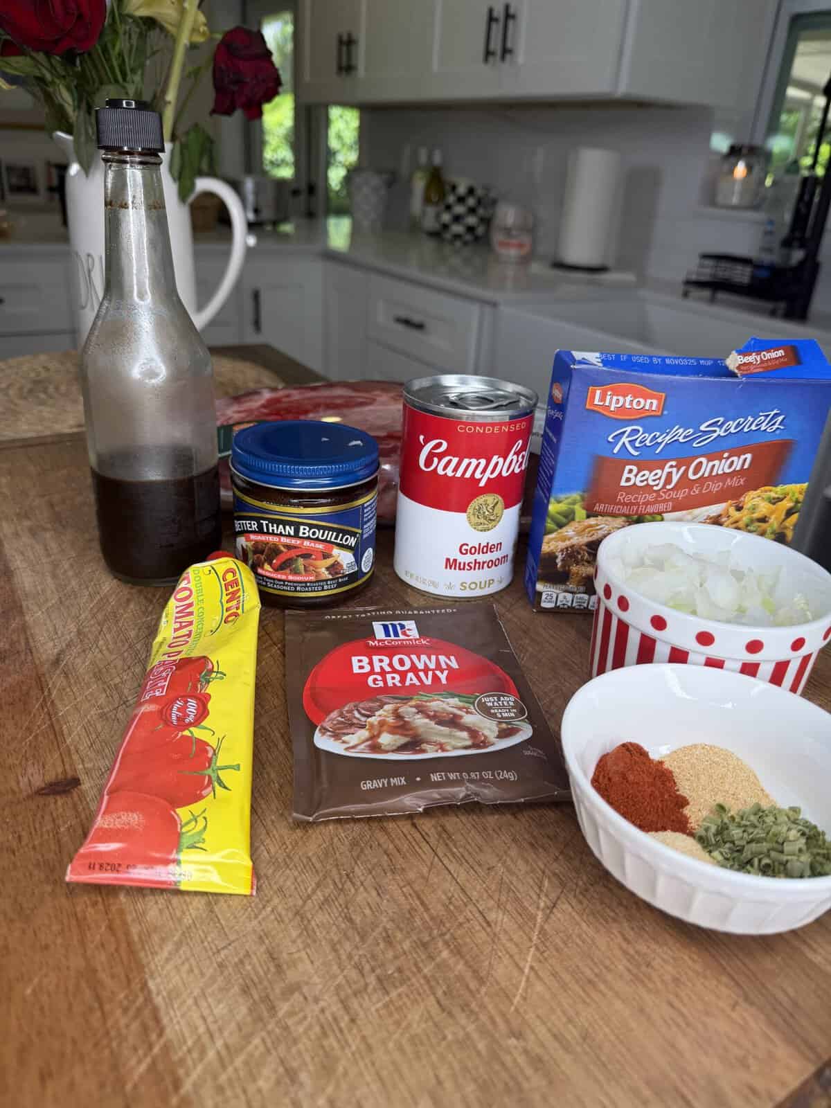 Ingredients on a wooden table: soy sauce, beef bouillon, golden mushroom soup, recipe mix, chopped onions, tomato paste, brown gravy mix, and a spice blend in a white bowl. Kitchen background.