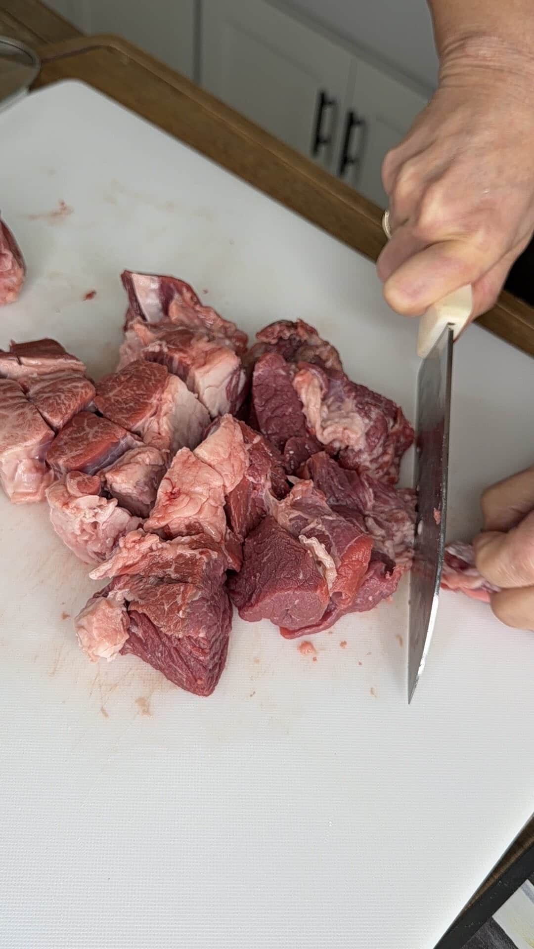 A person is cutting raw beef into chunks on a white cutting board with a large knife.