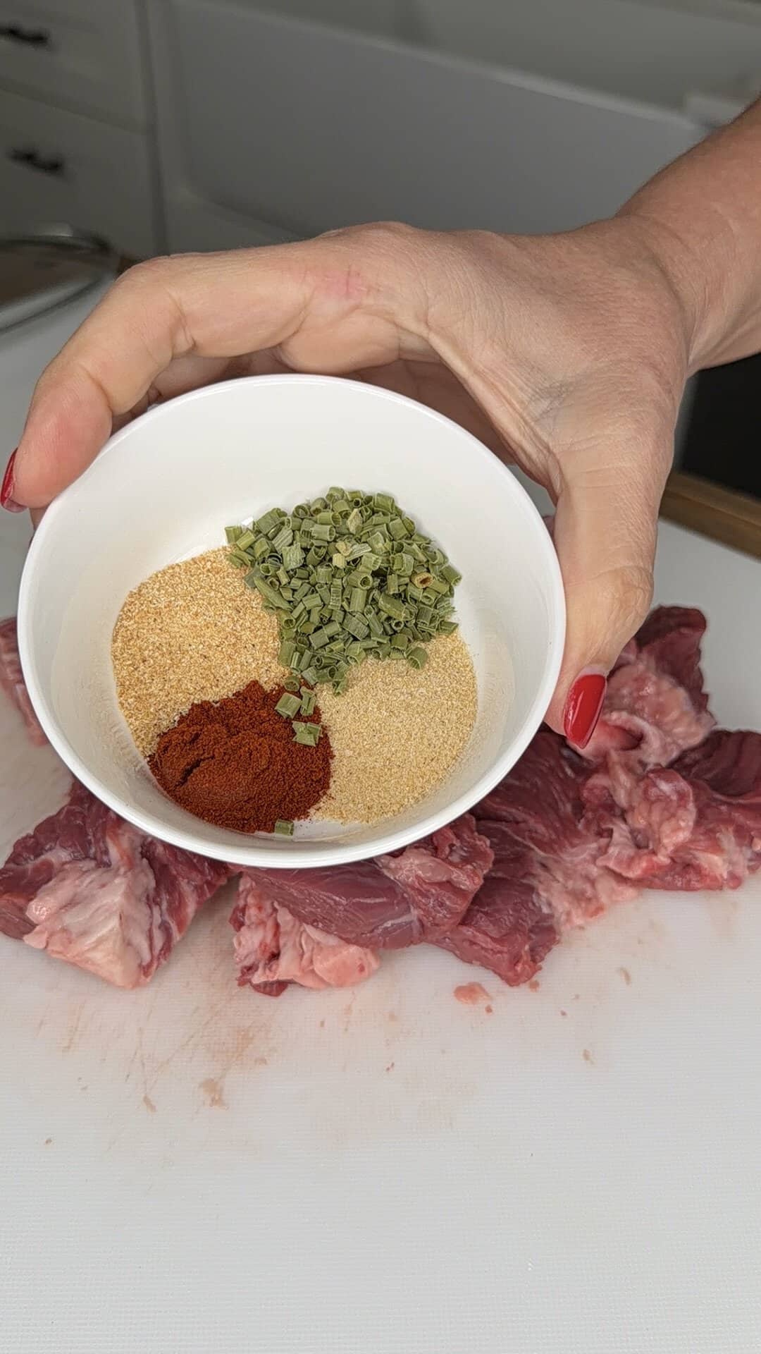 A hand with red nails holds a white bowl with dried chives, paprika, and garlic powder above raw pieces of beef on a white cutting board.