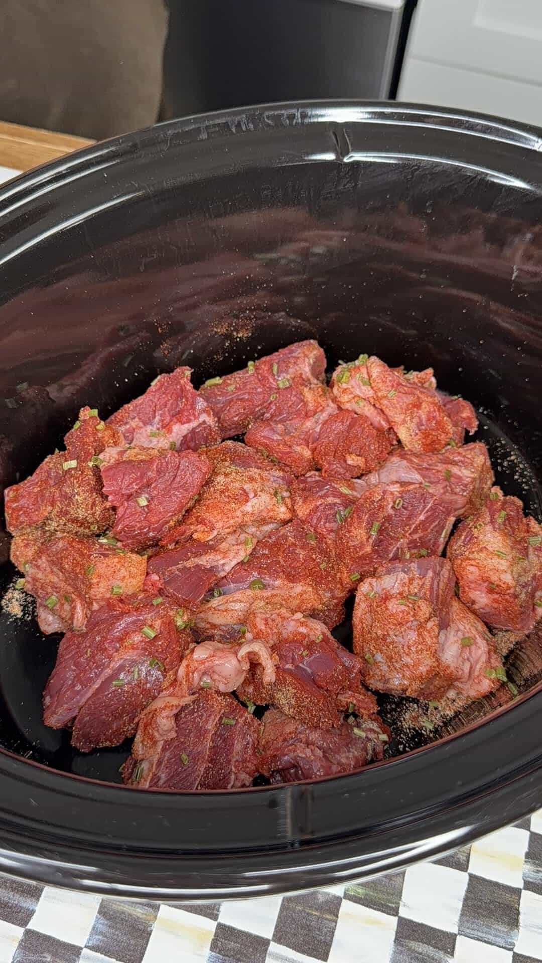 Chunks of raw beef seasoned with spices and green herbs sit in a black slow cooker, ready for cooking. The background shows a countertop with a checkered pattern and a kitchen appliance.
